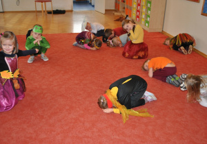 Dzieci klęczą lub kucają na dywanie zwijając się ciasno.